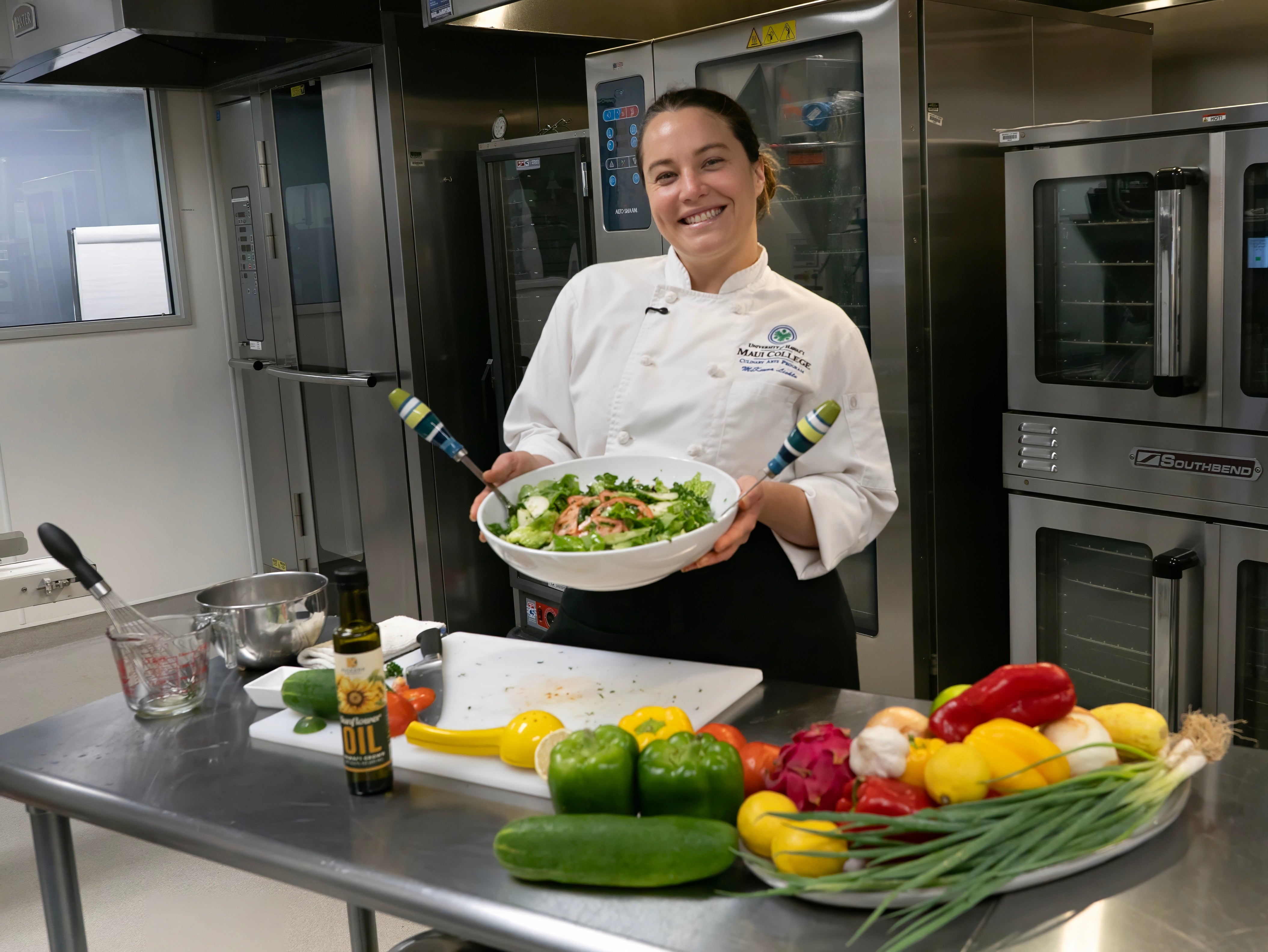 Chef holding a dish of salad in a kitchen with various vegetables and cooking equipment.